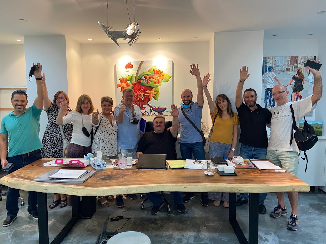 A cheerful group of ten colleagues gathers around a large live-edge wooden conference table in a bright, modern office space. The central colleague sits smiling behind a laptop and office documents, surrounded by teammates standing on either side who enthusiastically raise their hands, wave, and give thumbs-up gestures toward the camera. Colorful contemporary art pieces adorn the white walls in the background, completing the lively team atmosphere.