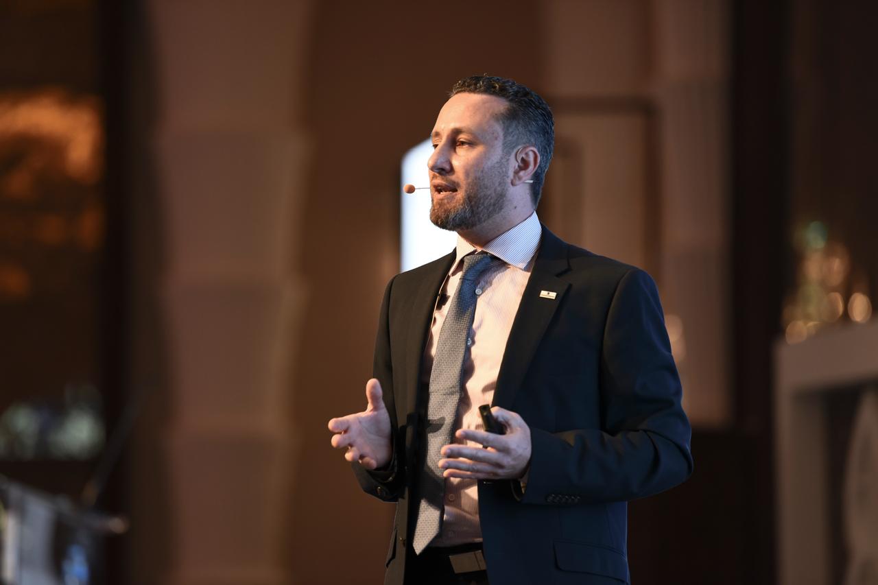 Makram Hani, CEO of Arms McGregor International and founder of Hype Agency Dubai Makram Hani speaking on stage during a business presentation or conference. He is wearing a tailored dark suit, light dress shirt, and patterned tie with a headset microphone, gesturing with his hands while addressing the audience against a softly lit background.