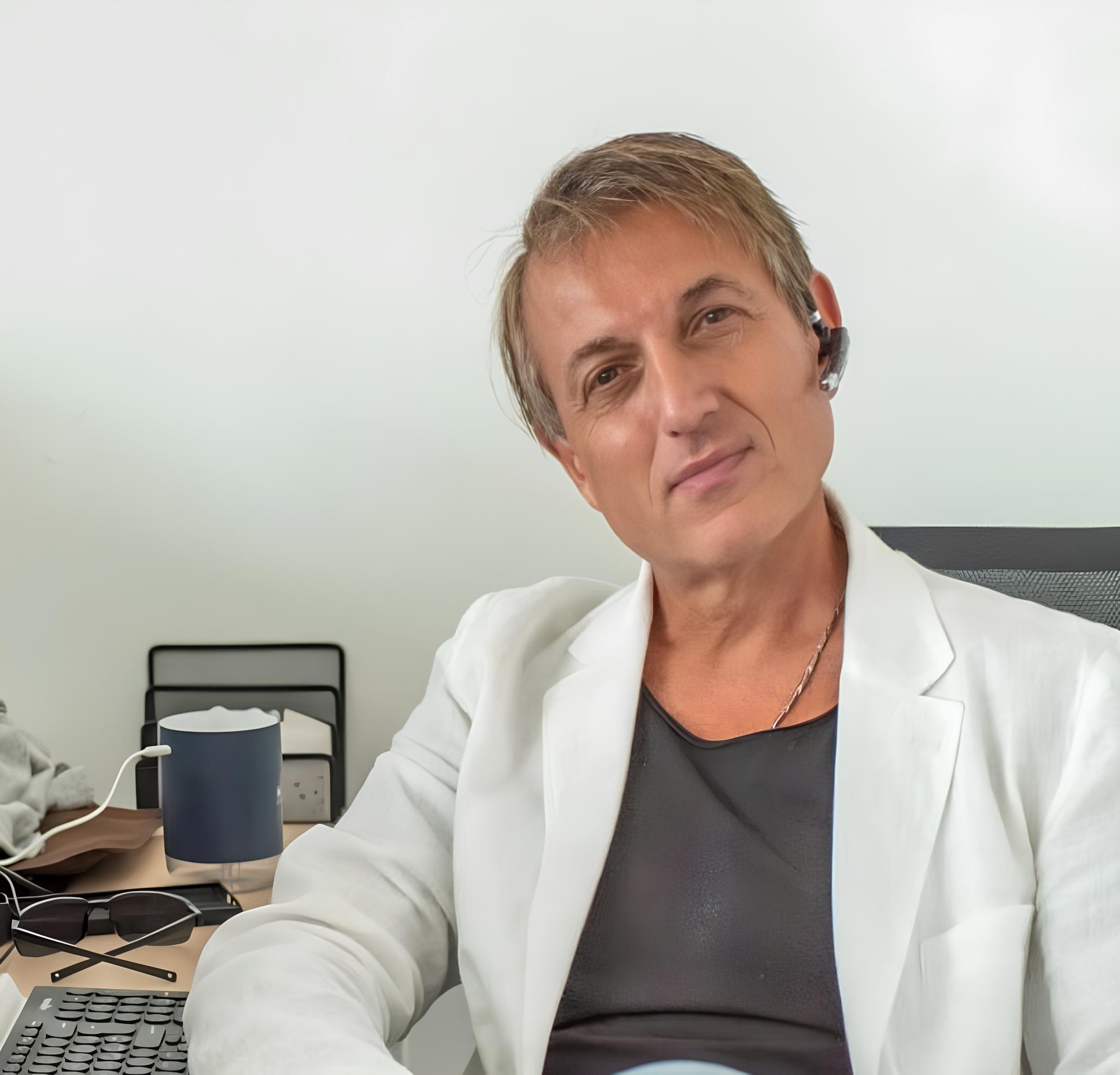 Andrea Giudice, SEO lead at Hype Agency Dubai and PrincipioAttivo A close-up portrait of Andrea Giudice seated at his office desk in a professional setting. He is wearing a white blazer over a dark top, a subtle necklace, and a wireless earpiece. The background shows an office workspace featuring a keyboard, mug, sunglasses, and neutral walls.